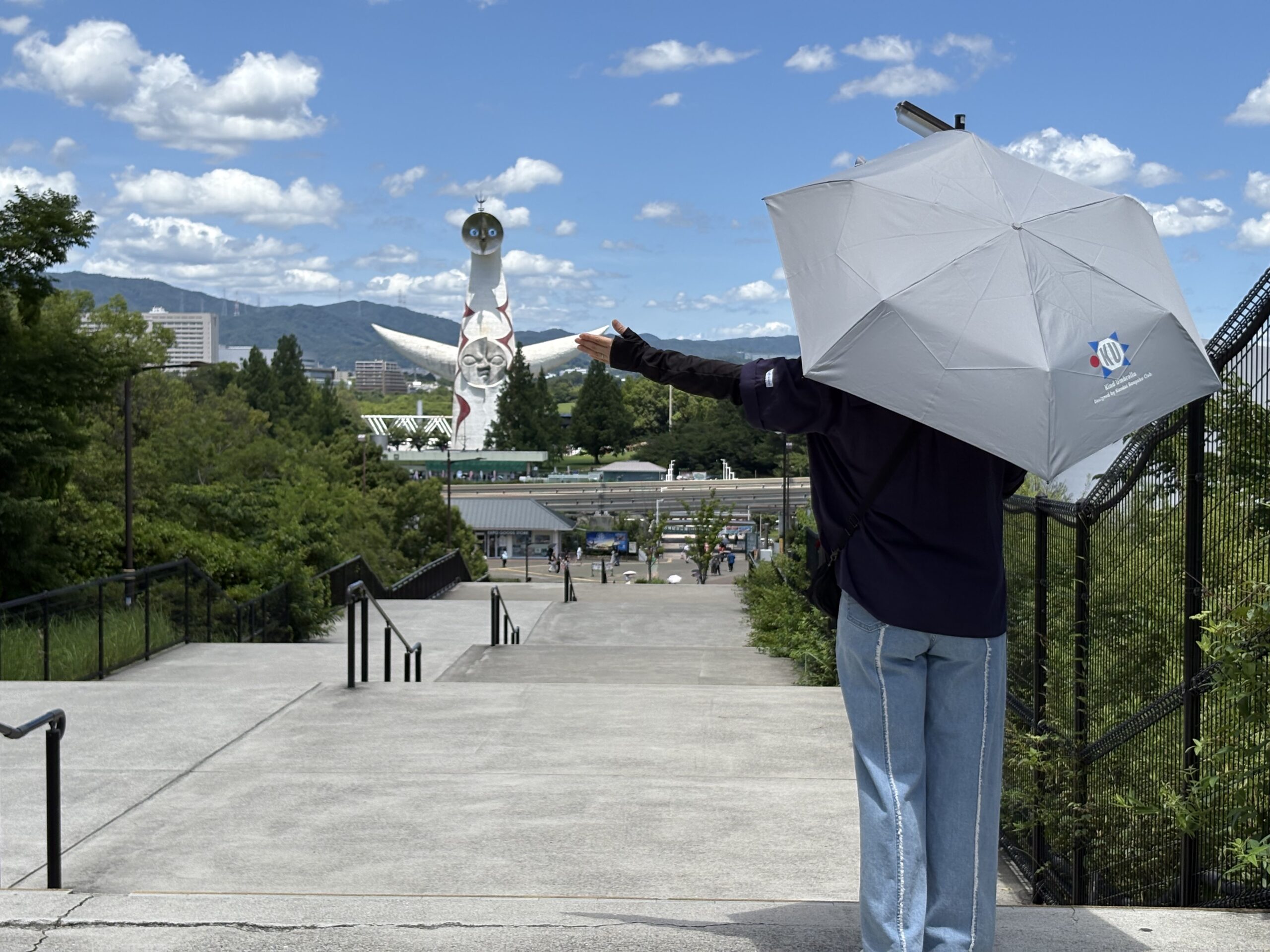 千里の万博記念公園中央口から数百メートル手前の階段で、太陽の塔と手をつなぐように片手を横に広げて関大万博部の晴雨兼用日傘をさす後ろ姿の女性の写真