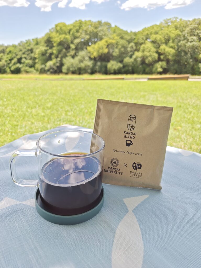 千里の万博記念公園内の芝生で、夏の陽ざしの中、木洩れ日に包まれて、ベンチに北欧風の白いお魚柄ランチョンマットを敷き、ガラス容器に淹れたコーヒーと、そのパッケージが飾られています