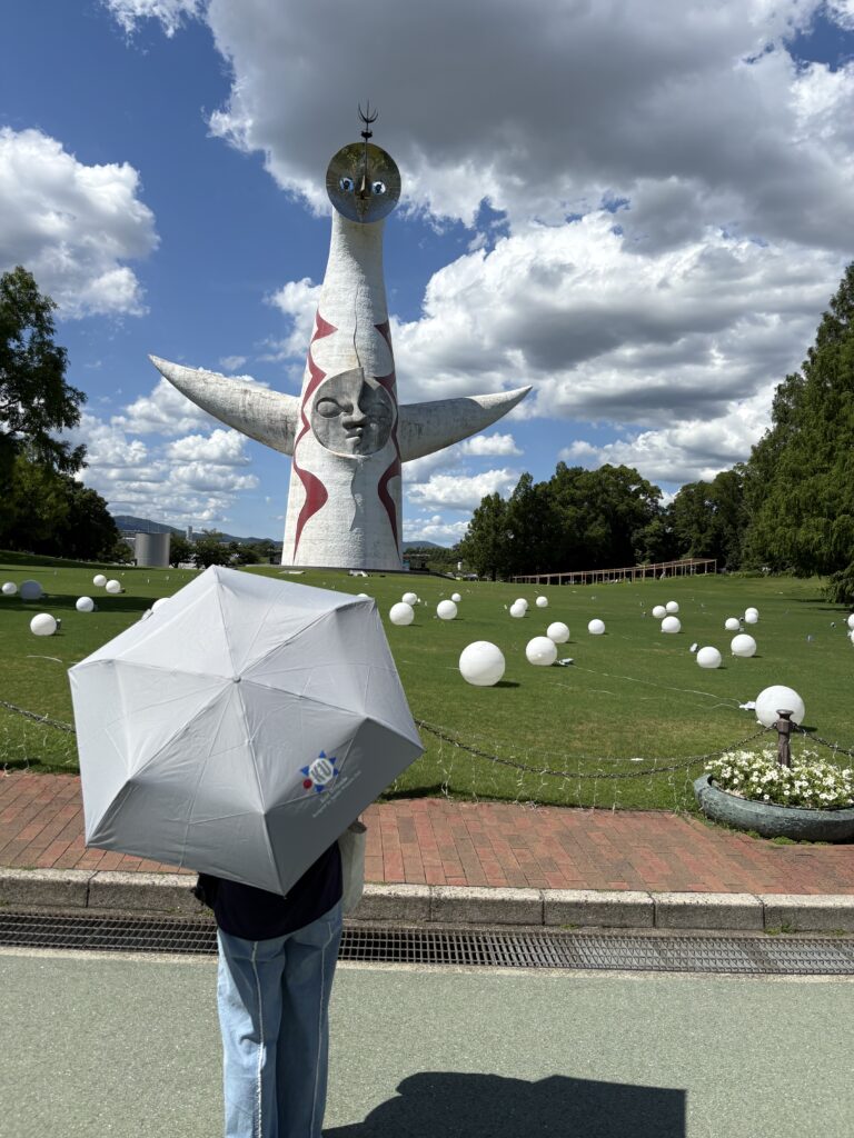 千里の万博記念公園内の太陽の塔の前で関大万博部がデザインした晴雨兼用傘をさして立っている女性の後ろ姿