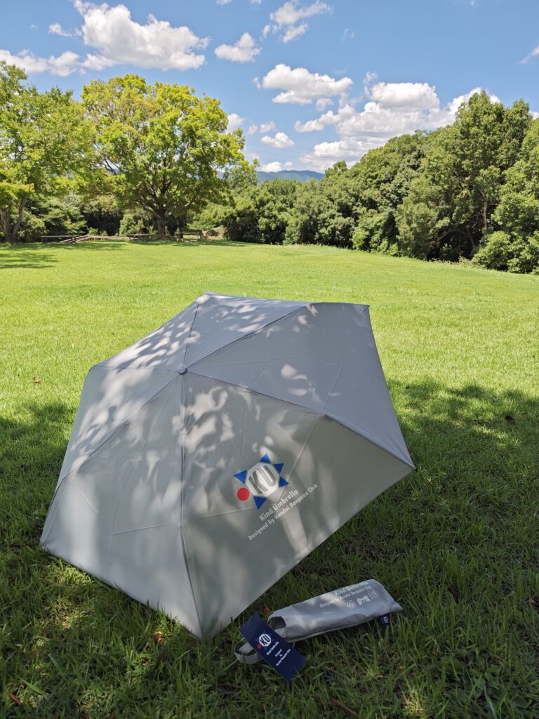 千里の万博記念公園内の花の丘の芝生の上で関大万博部がデザインした晴雨兼用傘を広げると傘に木洩れ日がうつる