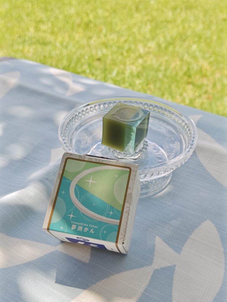 千里の万博記念公園内の芝生で、夏の陽ざしの中、木洩れ日に包まれて、ベンチに北欧風の白いお魚柄ランチョンマットを敷き、涼やかなガラス器に盛りつけられた「夢洲かん」。手前には箱が添えられています