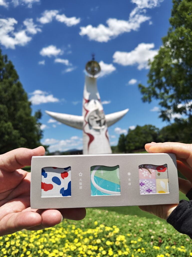 千里万博記念公園の太陽の塔の前で夏ギフト期間限定販売しているキューブ型ようかん「大阪ええYOKAN」3個入りの箱を2人が持ち上げている写真