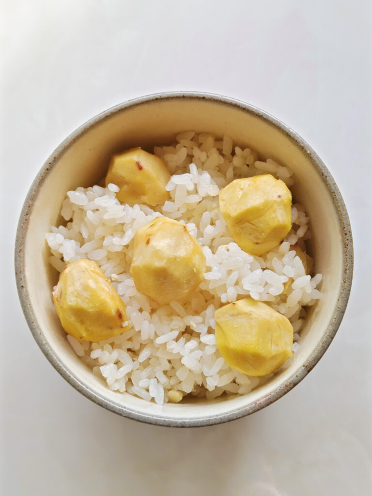 ベージュ色のお茶碗に、炊き上がった栗ごはんを盛りつけています。白ごはんの上に、一粒を半分にカットした栗を5～6個、のせています。栗の切り口は下にしています。