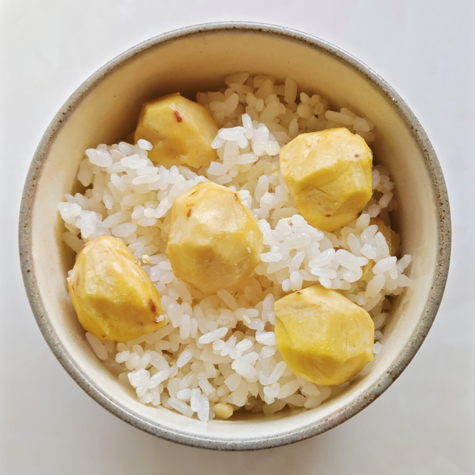 ベージュ色のお茶碗に、炊き上がった栗ごはんを盛りつけています。白ごはんの上に、一粒を半分にカットした栗を5～6個、のせています。栗の切り口は下にしています。