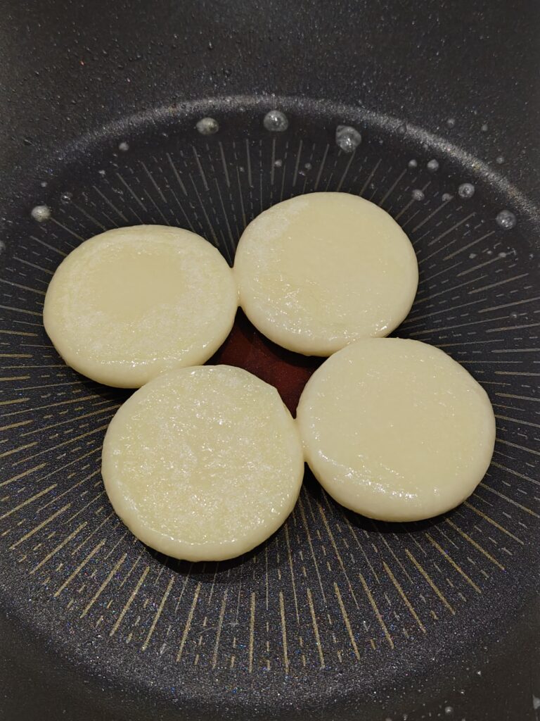 フライパンに丸もちを４つ並べています。丸もちは、地域特産「紅せんり」ブランド。丹波青垣産の滋賀羽二重もち使用「紅せんりのおもち」。もちピザを作るために、片面蒸し焼きしたおもちを裏返した写真です。