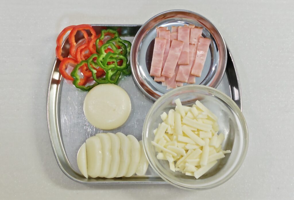 おもちピザの材料を並べています。丸もち２個。うすくカットしたピーマン、細く切ったベーコン、ピザ用のチーズ。