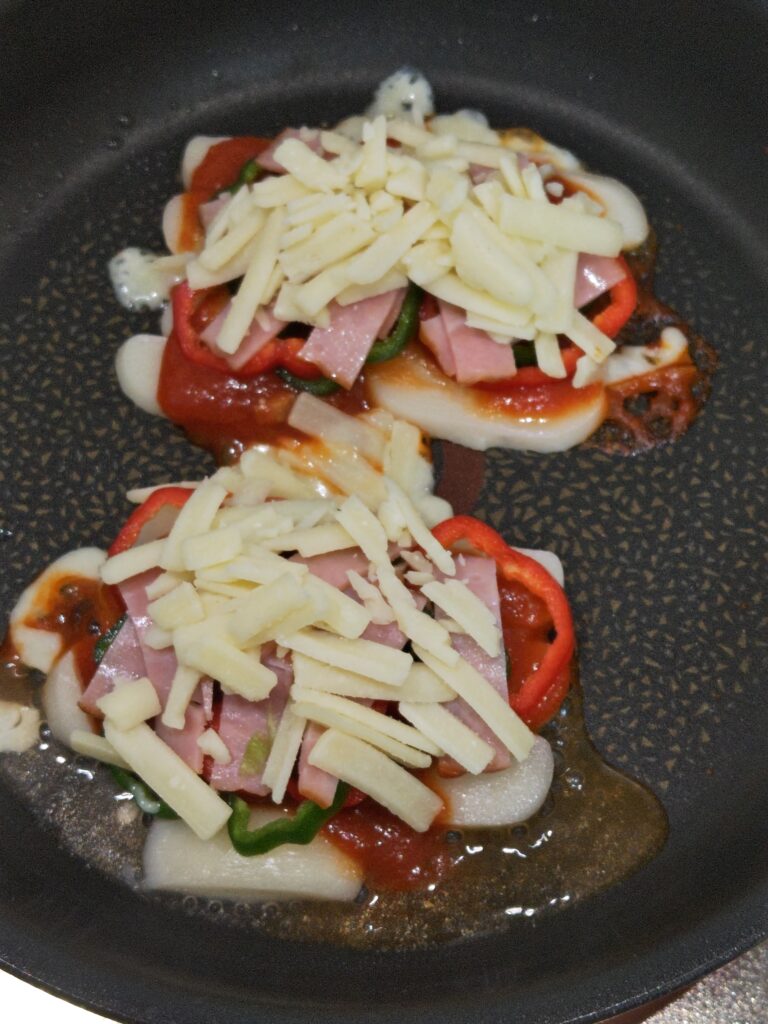 丸もちピザをフライパンで弱火で蒸し焼きしています。トッピングは、ピザソース、ベーコン、ピーマン、とろけるチーズです。