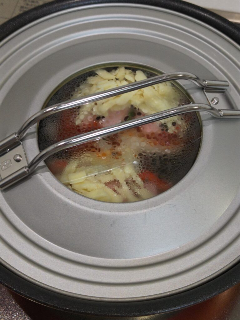 丸もちピザをフライパンで弱火で蒸し焼きしています。ピザソースをぬり、ベーコン、ピーマン、とろけるチーズをトッピングしたあと、フタをして蒸し焼きしています。