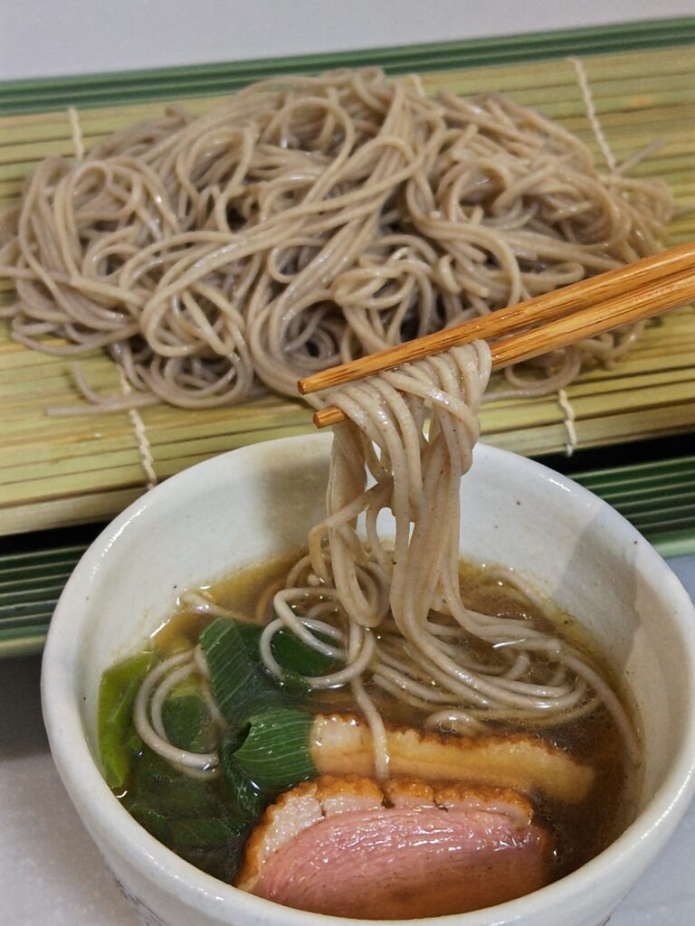 手前の白い和の器に、鴨ネギと蕎麦つゆが入っており、蕎麦の麺の一部をお箸で持ち上げています。蕎麦は関西大学と企業の産学連携で誕生した「九・九割蕎麦」です。