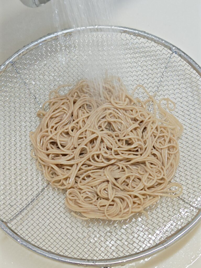 キッチンのシンクにザルを置いて、茹でたお蕎麦をお湯ごと流しいれ、蛇口から水をザーっと流して洗っています。