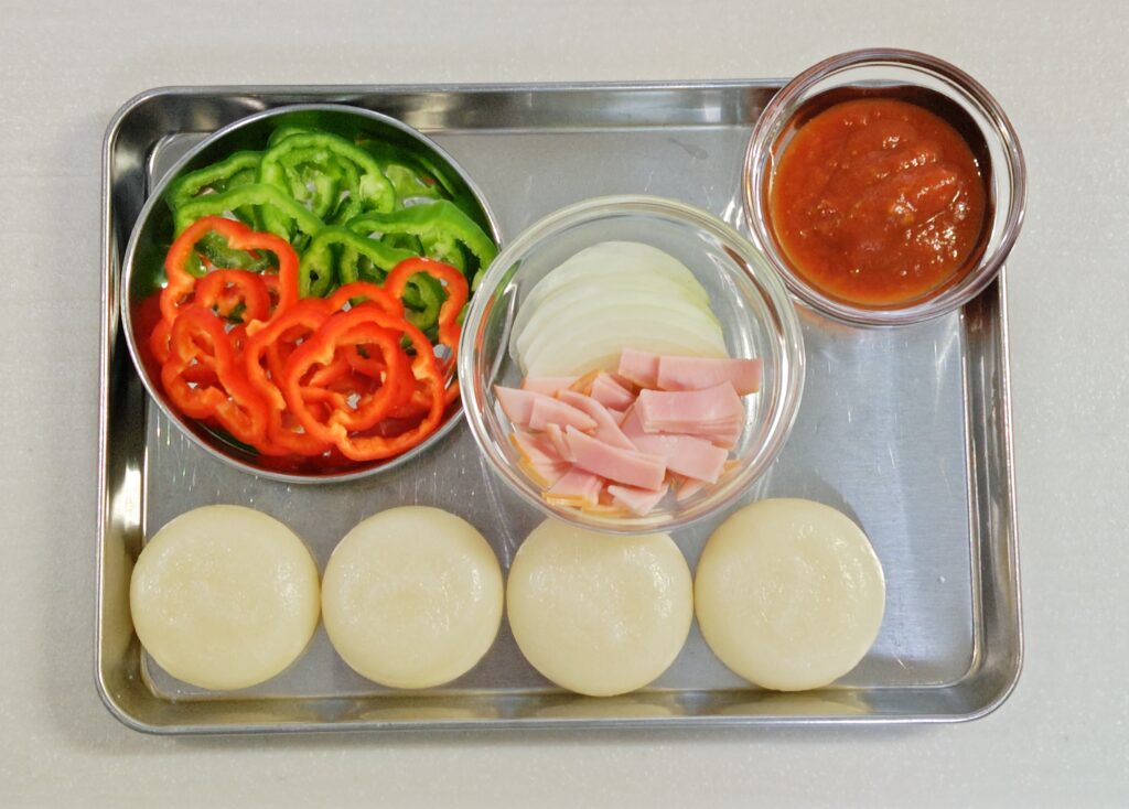 もちピザの材料が並んでいます。丸もち４個。輪切りした赤と緑のピーマン、うすくスライスした玉ねぎ、細くカットしたベーコン、ピザソース。