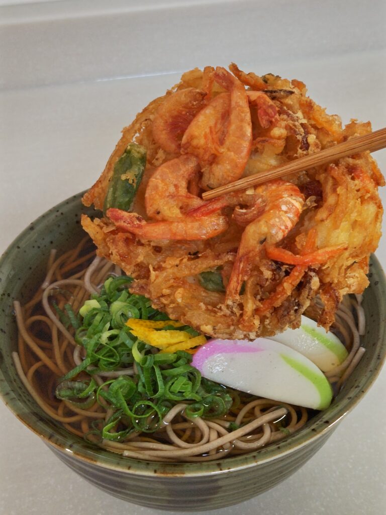 緑色の和の器に、蕎麦と、スライスしたかまぼこ、小口ネギ、柚の皮が入っており、お箸でつかんだかき揚げを器に盛り付けようとしています。蕎麦は関西大学と企業の産学連携で誕生した「九・九割蕎麦」です。