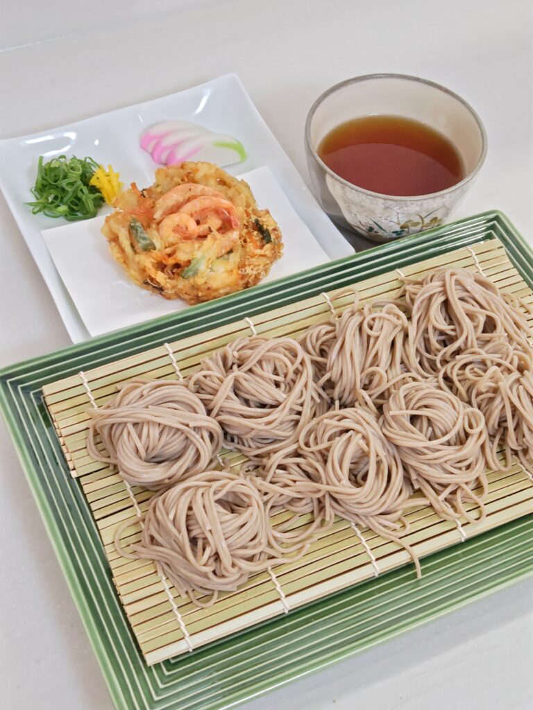 手前の緑色の四角い和の器に、竹ザルを敷き、蕎麦を小口で8つ盛りつけています。奥の白いお皿には、トッピングのかき揚げ、小口ネギ、スライスしたかまぼこ、柚の皮を盛りつけています。右隣に、丸い小さな器に蕎麦のつゆが入っています。蕎麦は、関西大学と企業の産学連携で誕生した「九・九割蕎麦」です。